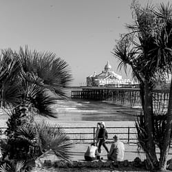 A pictorial black and white photo of Eastbourne pier with some people in the foreground viewed between trees